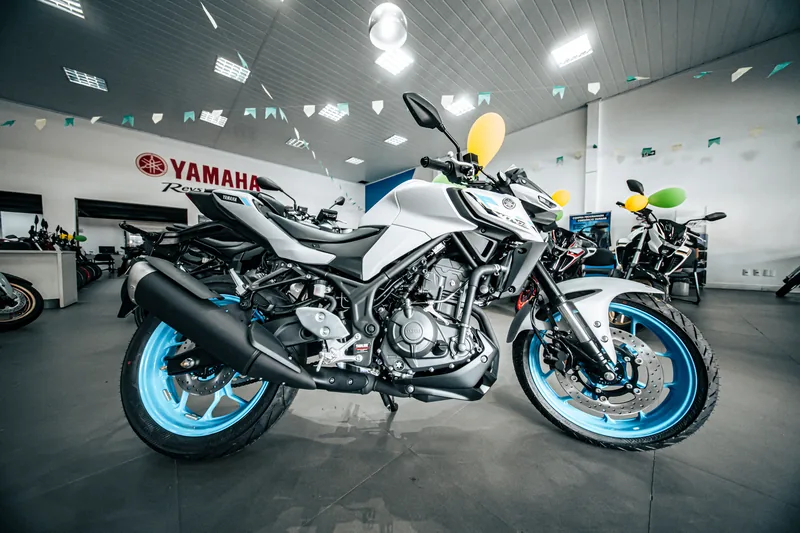 MT-03 branca com rodas azul-claras, vista de lado no salão da loja, com a parede Yamaha ao fundo.
